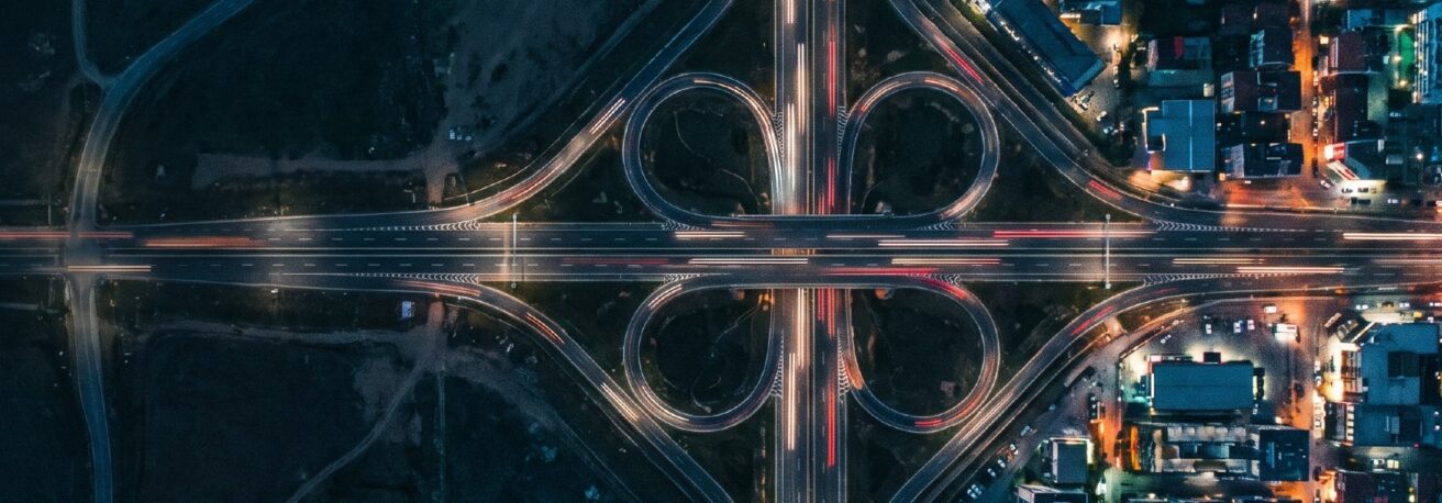 Aerial view of a motorway interchange at night, light trails on every ramp, the network an expedited run drives straight past